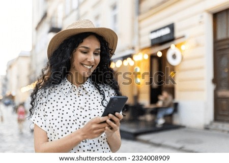 A beautiful young woman walks through the evening city in a hat, a smiling Latin American woman holds a smartphone in her hands. A tourist with curly hair types a message and browses online pages on