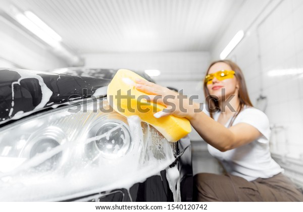 Beautiful young woman wash foam with yellow sponge\
headlights of car.
