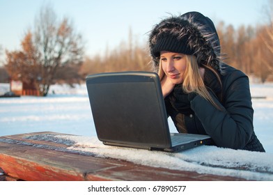 Beautiful young woman watching laptop sitting on a bench in winter surrounded by snow
