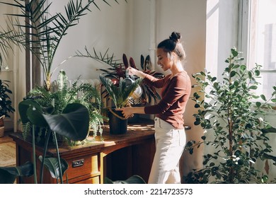 Beautiful young woman watering houseplants and smiling while standing at the domestic room - Powered by Shutterstock