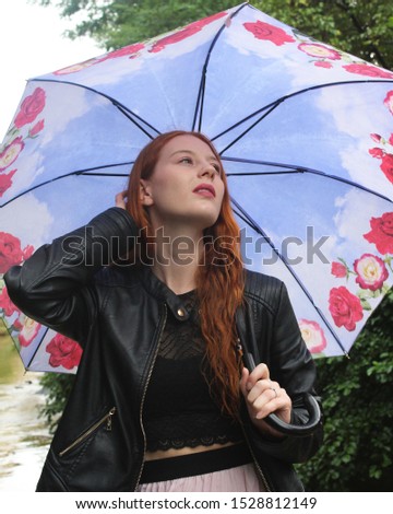 Beautiful young woman wearing a black leather jacket and holding a light blue umbrella with red rose designs on a rainy autumn day; vertical mid length.