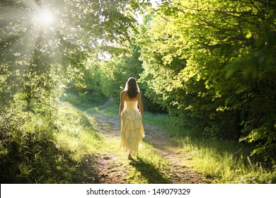 Hermosa joven mujer vestida con elegante vestido blanco caminando por un sendero forestal con rayos de luz solar que resplandece a través de las hojas de los árboles