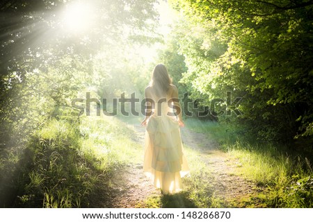 Beautiful young woman wearing elegant white dress standing with a smile on a road in the forest with rays of sunlight beaming through the leaves of the trees