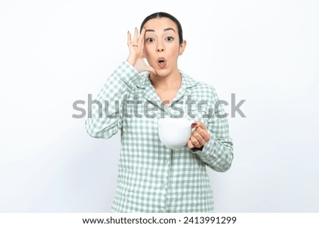 Beautiful young woman wearing green plaid pajama with scared expression, keeps hands on head, jaw dropped, has terrific expression. Omg concept