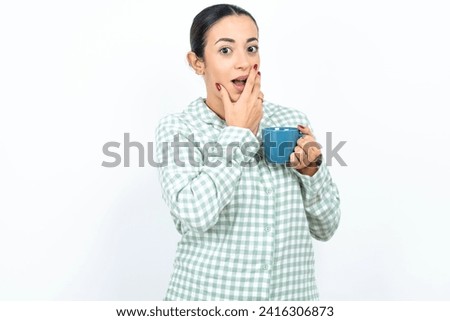 Beautiful young woman wearing green plaid pajama covers mouth and looks with wonder at camera, cannot believe unexpected rumors.