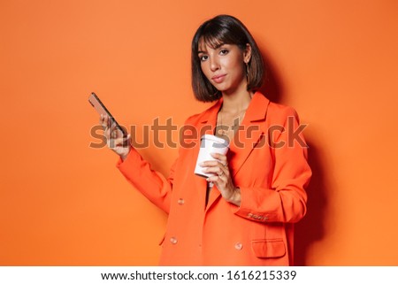 Beautiful young woman wearing jacket standing isolated over orange background, using mobile phone while drinking takeaway coffee