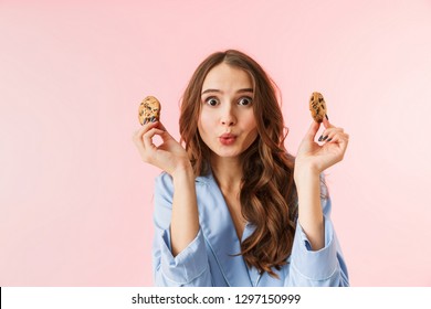 Beautiful Young Woman Wearing Pajamas Standing Isolated Over Pink Background, Eating Chocolate Cookies