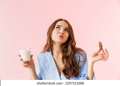 Beautiful young woman wearing pajamas standing isolated over pink background, eating chocolate cookies, drinking milk