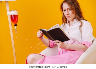 Beautiful Young Woman Wearing Patient Clothes Sitting In Low Chair, Taking Her Medicine From Infusion Drip Bottle While Reading A Book. Over Orange Background