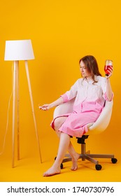 Beautiful Young Woman Wearing Patient Clothes Sitting In Low Chair, Taking Her Medicine From Infusion Drip Bottle. Over Orange Background