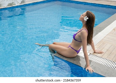 Beautiful Young Woman Wearing Purple Bikini Bathing Suit, Dipping Her Feet In The Pool Water. Female Model With Plumeria Flower In Her Wet Blonde Hair. Background, Copy Space, Close Up