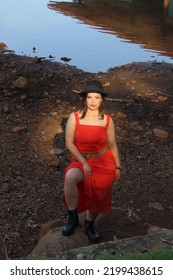 Beautiful Young Woman Wearing Small Black Hat, Red Dress And Black Boots, Standing, Viewed From Above, In A Beautiful Park