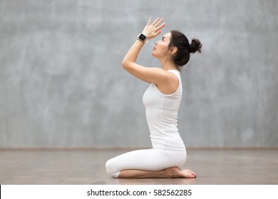 Beautiful young woman wearing sport watch for heart rate and calorie burn control working out against grey wall, doing yoga or pilates exercise. Girl sitting in seiza, vajrasana pose. Full length 