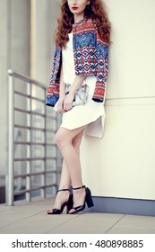 Beautiful Young Woman Wearing A White Dress And A Bomber Jacket, Holding A Silver Clutch Bag