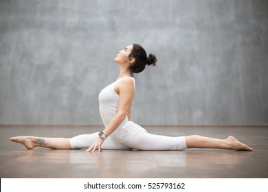 Beautiful young woman wearing white sportswear set and smartwatch working out against grey wall, doing yoga or pilates exercise. Monkey god, splits, hanumanasana. Full length