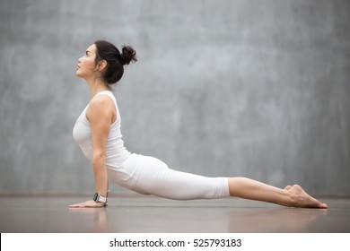 Beautiful young woman wearing white sportswear set and smartwatch working out against grey wall, doing yoga or pilates exercise. Upward facing dog pose, Urdhva mukha shvanasana. Full length