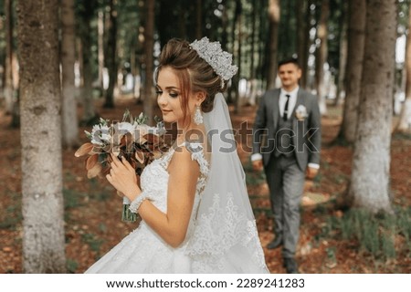 A beautiful young woman in a wedding dress between tall trees in the forest with a royal hairstyle and a chic tiara with a bouquet of flowers in her hands, a wedding in golden color