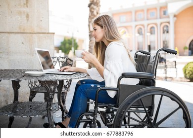 Beautiful Young Woman In A Wheelchair Doing Some Work On Her Laptop And Enjoying A Cup Of Coffee In A Restaurant