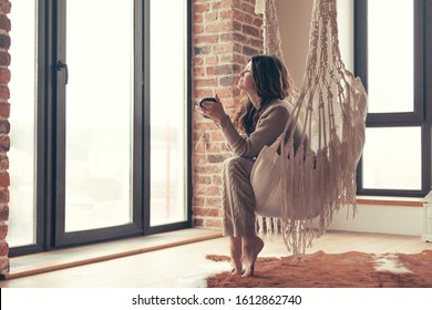 Beautiful young woman, which is wearing cashmere nightwear clothes, drinking tea or coffee while relaxing on rocking chair in scandinavian cabin near big window one lazy weekend morning