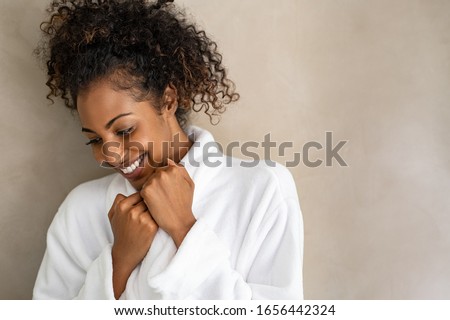 Beautiful young woman in a white bath robe isolated on beige background with copy space. Happy african girl in bathrobe against light brown wall. African american woman enjoy after body treatment.
