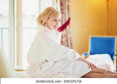 Beautiful Young Woman In White Bathrobe Sitting On Bed At Early Morning And Stretching After Night Sleep