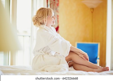 Beautiful Young Woman In White Bathrobe Sitting On Bed At Early Morning And Stretching After Night Sleep