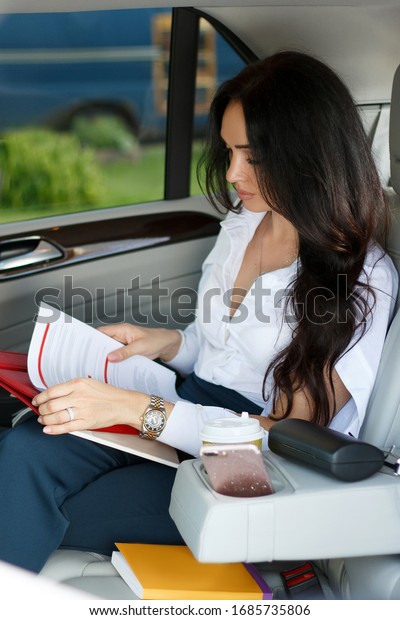 Beautiful young woman in\
white blouse and black trousers sits in the back seat of her car\
and reads books and notebooks. Business. Distance Learning Concept.\
E-learning. 