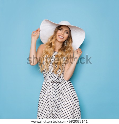 Beautiful young woman in white dotted summer dress and sun hat is smiling and looking at camera. Three quarter length studio shot on turquoise background.