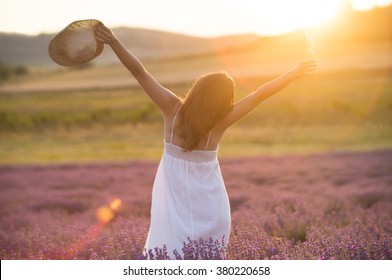 Beautiful young woman with a white dress and a straw hat standing in the middle of a lavender field at in the golden light of the sunset praising the beauty of life.