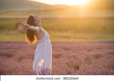 Beautiful young woman with a white dress and a straw hat standing in the middle of a lavender field at in the golden light of the sunset praising the beauty of life.