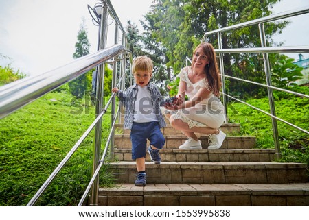 Beautiful young woman in a white dress and long hair with a cute little baby blonde boy at the green garden in summer, spring or autumn day
