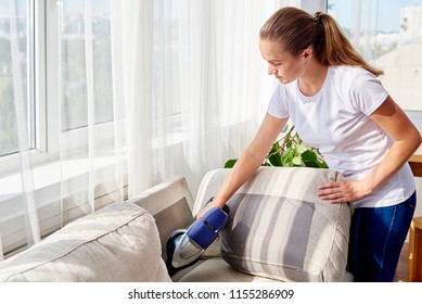 Beautiful young woman in white shirt and jeans cleaning sofa with vacuum cleaner in living room, copy space. Housework, spring-cleanig and chores concept. House hygiene, cleaning vacuum appliances 