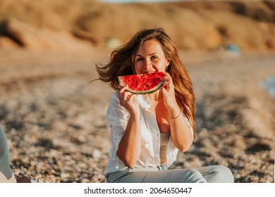 A beautiful young woman in a white shirt walks on the beach by the sea and eats watermelon at sunset. Quiet summer evenings on vacation