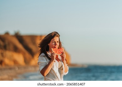 A beautiful young woman in a white shirt walks on the beach by the sea and eats watermelon at sunset. Quiet summer evenings on vacation