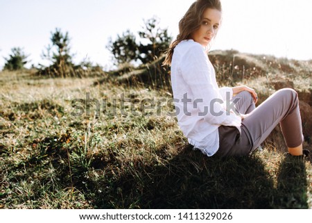 Beautiful young woman in white sits at the grass inrural sunny landscape background in summer. Tender happy woman in wild field enjoying nature. Back view