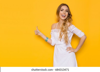 Beautiful young woman in white summer dress is showing thumb up, holding hand on hip, laughing and looking at camera. Three quarter length studio shot on yellow background. - Powered by Shutterstock