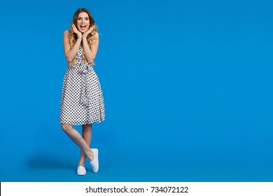 Beautiful Young Woman In White Summer Dress And Sneakers Is Holding Head In Hands, Shouting And Looking At Camera. Full Length Studio Shot On Blue Background.