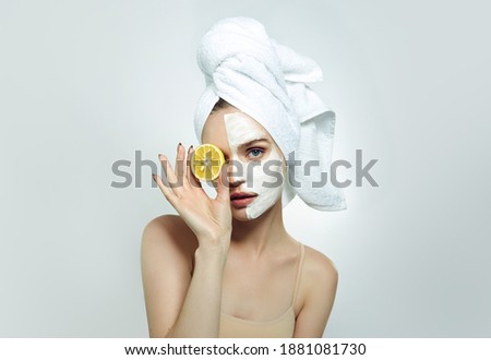 A beautiful young woman with a white towel on her head and a cosmetic cream mask on her face is holding a slice of lemon. Cosmetology.