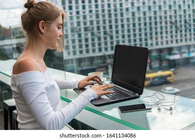 Beautiful young woman in white t-shirt is working on laptop and smiling while sitting outdoors in cafe. Young female using laptop for work. Female freelancer working on laptop in an outdoor cafe.