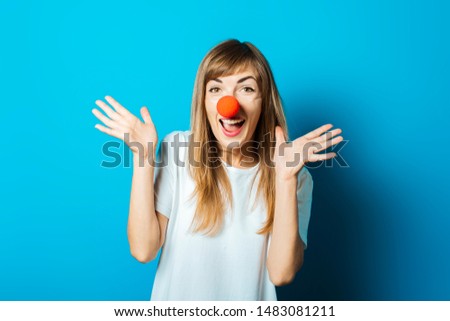 Beautiful young woman in a white T-shirt and a red clown nose smiles and makes hand gestures on a blue background. Concept party, costume, red nose day