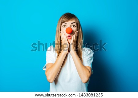 Beautiful young woman in a white T-shirt and red nose of a clown with a surprised face on a blue background. Concept party, costume, red nose day