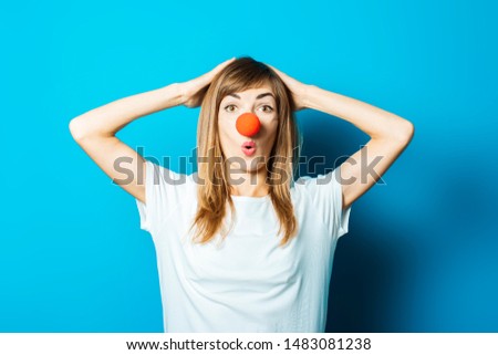 Beautiful young woman in a white T-shirt and red nose of a clown with a surprised face on a blue background. Concept party, costume, red nose day