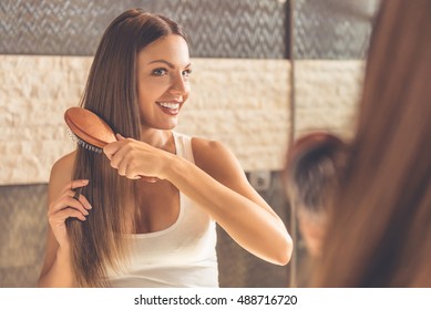 Beautiful young woman in white undershirt is combing her hair and smiling while looking into the mirror in bathroom - Powered by Shutterstock