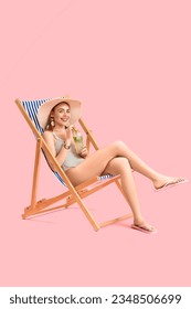 Beautiful young woman in wicker hat with glass of tasty mojito sitting on deckchair against pink background