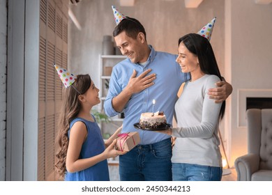 Beautiful young woman wife and her cute daughter girl congratulating father with birthday wearing party hats. Woman standing with holiday cake in hand, little girl with present box for daddy father - Powered by Shutterstock