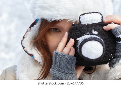 Beautiful young woman in winter clothing, fingerless mittens and hat with white fur photographing by a black knitted camera. Shallow dof. Focus on eye.