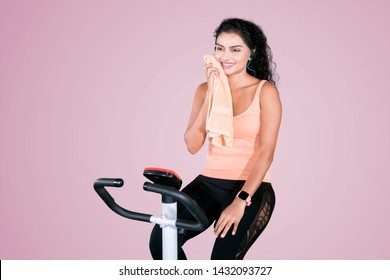 Beautiful Young Woman Wiping Her Sweat With A Towel After Workout On An Exercise Bike With Purple Background