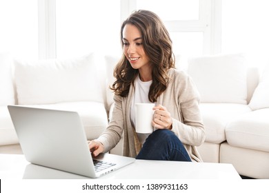 Beautiful young woman working on laptop computer while sitting at the living room, drinking coffee - Shutterstock ID 1389931163