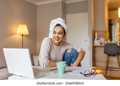 Beautiful young woman working on laptop computer while sitting at the living room, drinking coffee with face mask. Beautiful young woman smiling with face mask and looking at laptop screen - Powered by Shutterstock