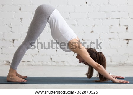 Beautiful young woman working out indoors, doing yoga exercise in the room with white walls, downward facing dog pose, adho mukha svanasana (sun salutation pose), full length, side view